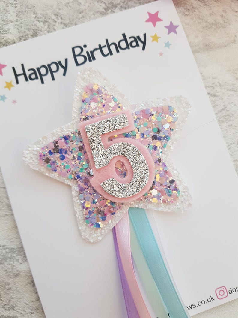 May include: A birthday card with the words "Happy Birthday" at the top. The card features a star-shaped decoration with the number 5 in silver glitter, surrounded by pink and iridescent glitter. Ribbons in pastel colors hang from the bottom.