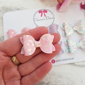 Peut inclure: Un ensemble de quatre n&oelig;uds pour cheveux en paillettes roses et bleues. Les n&oelig;uds sont dispos&eacute;s sur une carte blanche avec le texte "Donna's Bows" et un nom de profil sur les r&eacute;seaux sociaux.