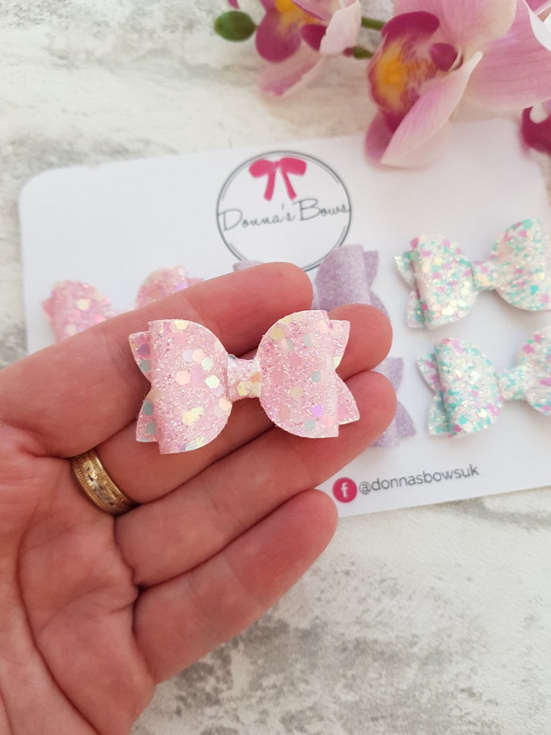 Peut inclure: Un ensemble de quatre n&oelig;uds pour cheveux en paillettes roses et bleues. Les n&oelig;uds sont dispos&eacute;s sur une carte blanche avec le texte "Donna's Bows" et un nom de profil sur les r&eacute;seaux sociaux.