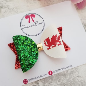 May include: A green and red glitter bow with a white bow featuring a red dragon. The bow is attached to a gold clip. The bow is displayed on a white card with the text "Donna's Bows" and social media handles.