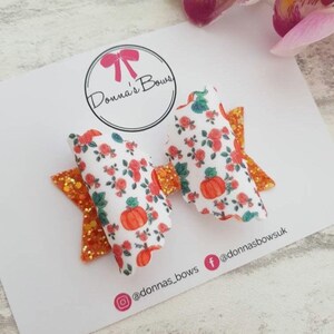 May include: A hair bow featuring a white fabric with a repeating pattern of orange pumpkins and small red flowers, accented by orange glitter fabric layers. The bow is attached to a white display card with the text Donna's Bows and social media handles.