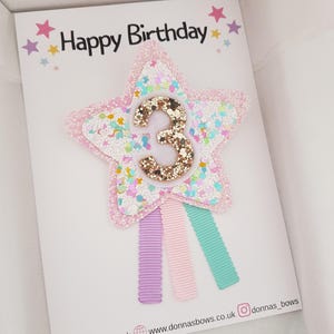 May include: A birthday card with a star-shaped hair clip. The star is pink with glitter and a gold number 3. The card says "Happy Birthday" with star accents. Three pastel-colored ribbons are attached to the star.