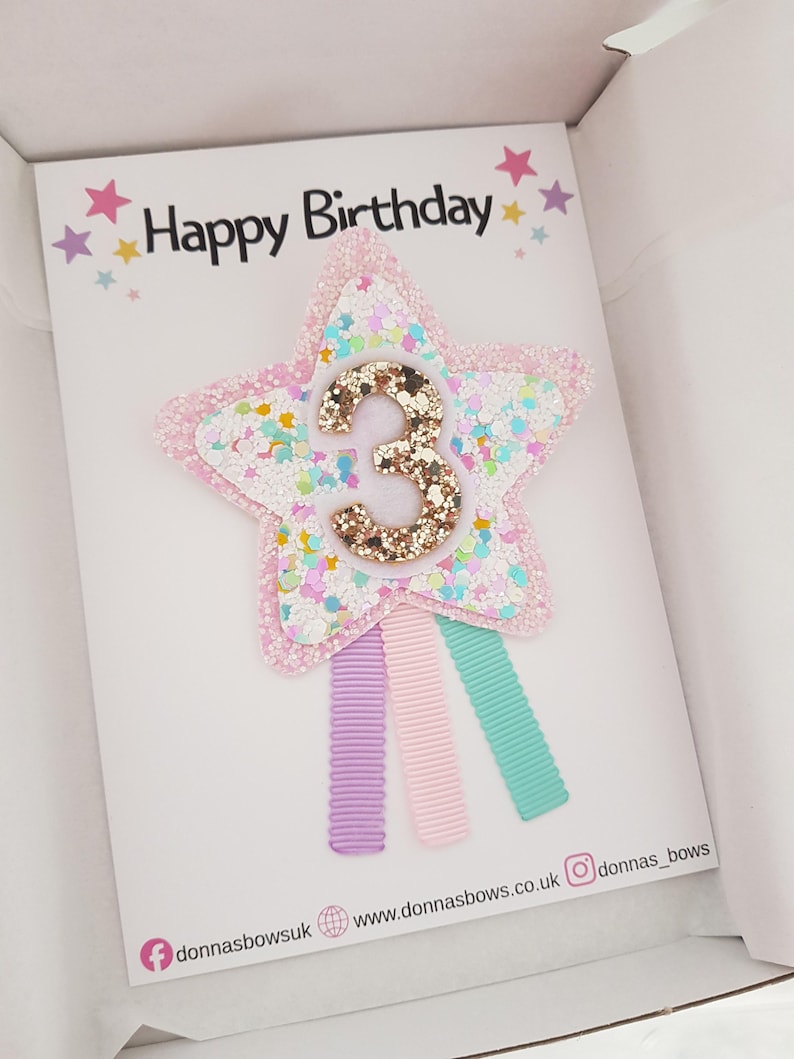 May include: A birthday card with a star-shaped hair clip. The star is pink with glitter and a gold number 3. The card says "Happy Birthday" with star accents. Three pastel-colored ribbons are attached to the star.