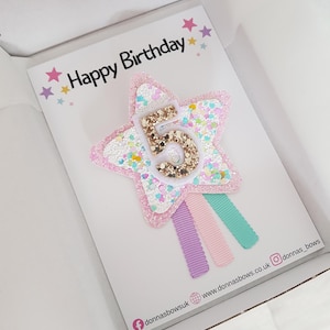 May include: A birthday card inside a white box. The card features a star-shaped hair clip with the number 5 in gold glitter. The star is pink with white and pastel-colored glitter accents. The card reads "Happy Birthday" with star decorations.