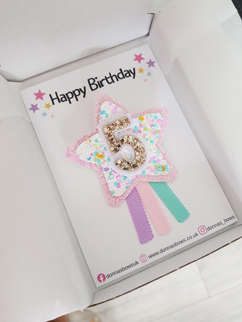 May include: A birthday card inside a white box. The card features a star-shaped hair clip with the number 5 in gold glitter. The star is pink with white and pastel-colored glitter accents. The card reads "Happy Birthday" with star decorations.