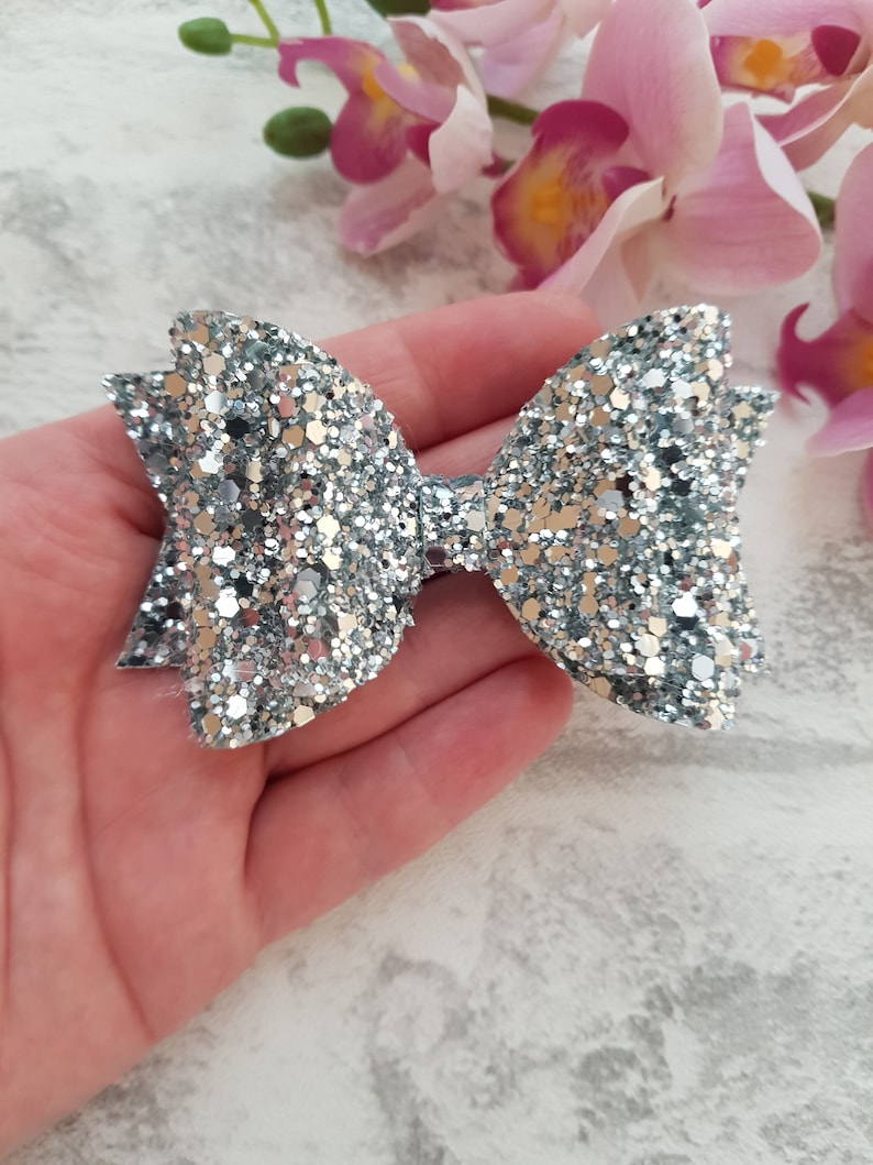 Peut inclure: Un n&oelig;ud en paillettes argent&eacute;es avec une finition scintillante. Le n&oelig;ud est fait de tissu et a une boucle &agrave; l'arri&egrave;re pour l'attacher aux cheveux.