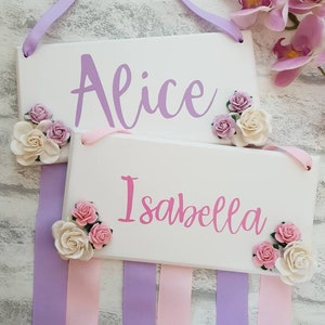 May include: Two rectangular white wooden signs featuring the names Alice and Isabella in purple and pink script lettering. Each sign is decorated with small artificial cream and pink roses and hangs from lavender and light pink fabric ribbons.