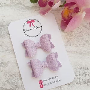 Peut inclure: Deux n&oelig;uds pour cheveux en paillettes lavande sur une carte blanche avec le texte "Donna's Bows" et les poign&eacute;es de m&eacute;dias sociaux @donnas_bows et @donnasbowsuk.