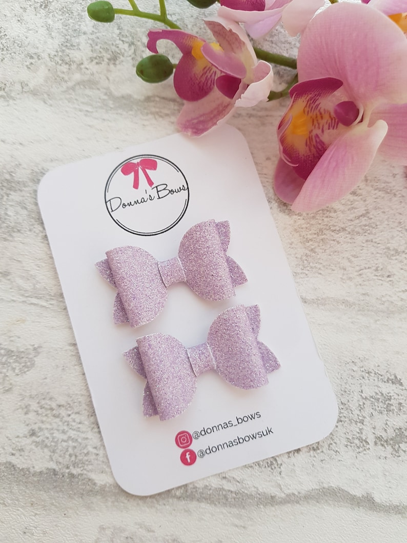 Peut inclure: Deux n&oelig;uds pour cheveux en paillettes lavande sur une carte blanche avec le texte "Donna's Bows" et les poign&eacute;es de m&eacute;dias sociaux @donnas_bows et @donnasbowsuk.