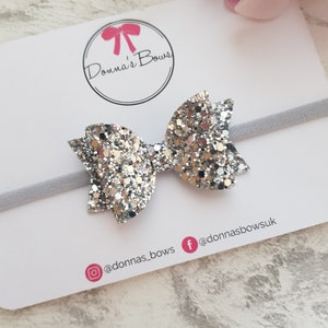 Peut inclure: Un serre-t&ecirc;te avec un n&oelig;ud en paillettes argent&eacute;es et un &eacute;lastique blanc. Le serre-t&ecirc;te est sur une carte blanche avec le texte "Donna's Bows" et les poign&eacute;es de m&eacute;dias sociaux @donnas_bows et @donnasbowsuk.