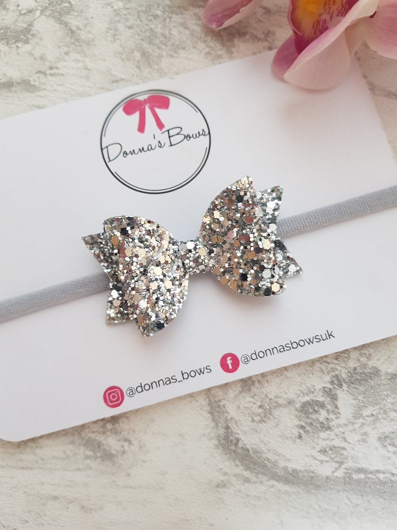 Peut inclure: Un serre-t&ecirc;te avec un n&oelig;ud en paillettes argent&eacute;es et un &eacute;lastique blanc. Le serre-t&ecirc;te est sur une carte blanche avec le texte "Donna's Bows" et les poign&eacute;es de m&eacute;dias sociaux @donnas_bows et @donnasbowsuk.