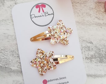May include: Two gold glitter star hair clips. The stars are a light gold color with pink and white glitter. The clips are on a white card with the text "Donna's Bows" and social media handles.