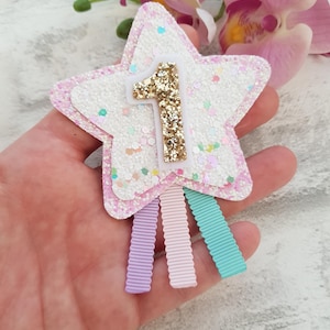 May include: A star-shaped hair clip with a glittery white and pink design. The number "1" is in gold glitter. Three pastel-colored ribbons are attached to the bottom. A festive accessory for a first birthday celebration.