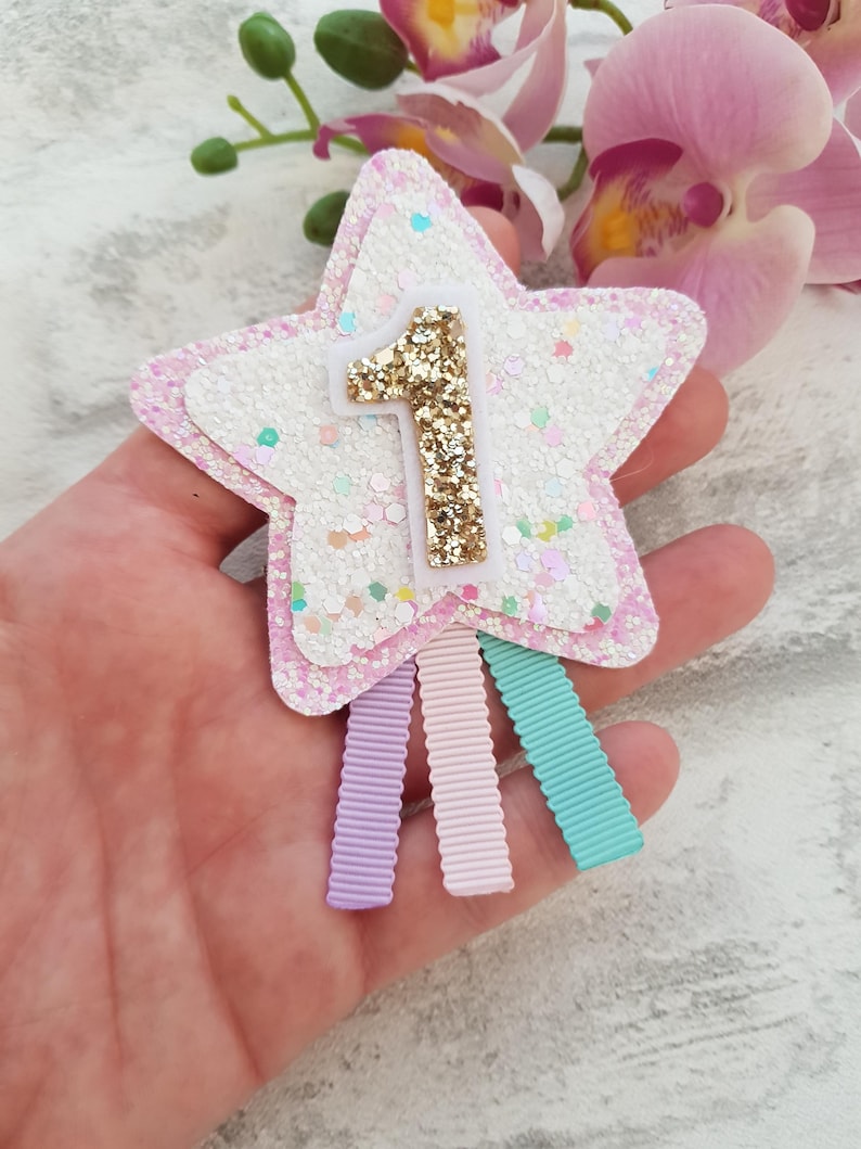 May include: A star-shaped hair clip with a glittery white and pink design. The number "1" is in gold glitter. Three pastel-colored ribbons are attached to the bottom. A festive accessory for a first birthday celebration.