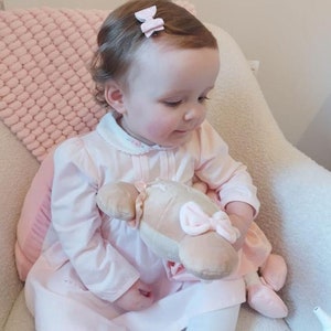 Peut inclure: Une petite fille portant une robe rose avec un col blanc et tenant un jouet en peluche. Elle a deux n&oelig;uds roses dans les cheveux.