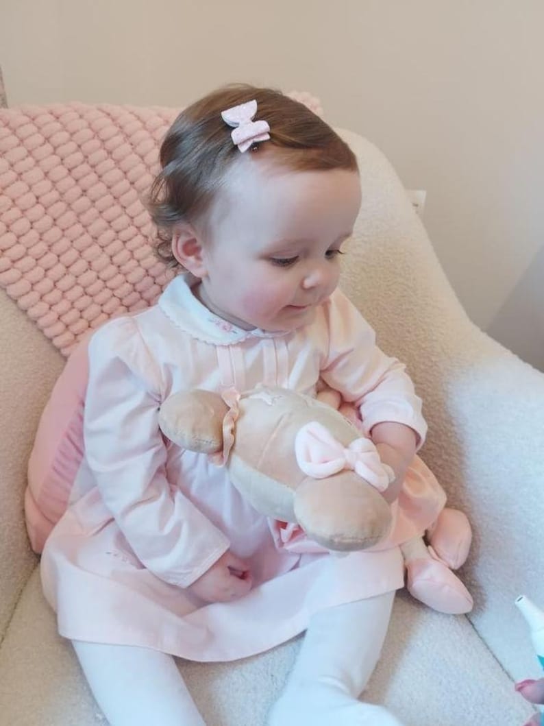 Peut inclure: Une petite fille portant une robe rose avec un col blanc et tenant un jouet en peluche. Elle a deux n&oelig;uds roses dans les cheveux.