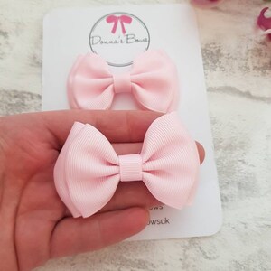 May include: Two light pink hair bows on a white card with the text "Donna's Bows". The bows are made of a ribbed fabric and are in a classic bow shape. The bows are small and delicate, perfect for a baby or young child.