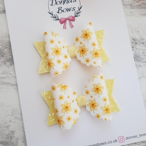 May include: Two hair bows with a daffodil pattern on a white background. The bows have a yellow glittery accent and are attached to a white card. The card has the text "Donna's Bows" and social media handles.