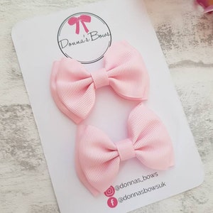May include: Two light pink ribbon hair bows on a white card. The card has a circular logo with a pink bow and the text "Donna's Bows". Social media handles are also visible. The bows are made of a ribbed fabric.