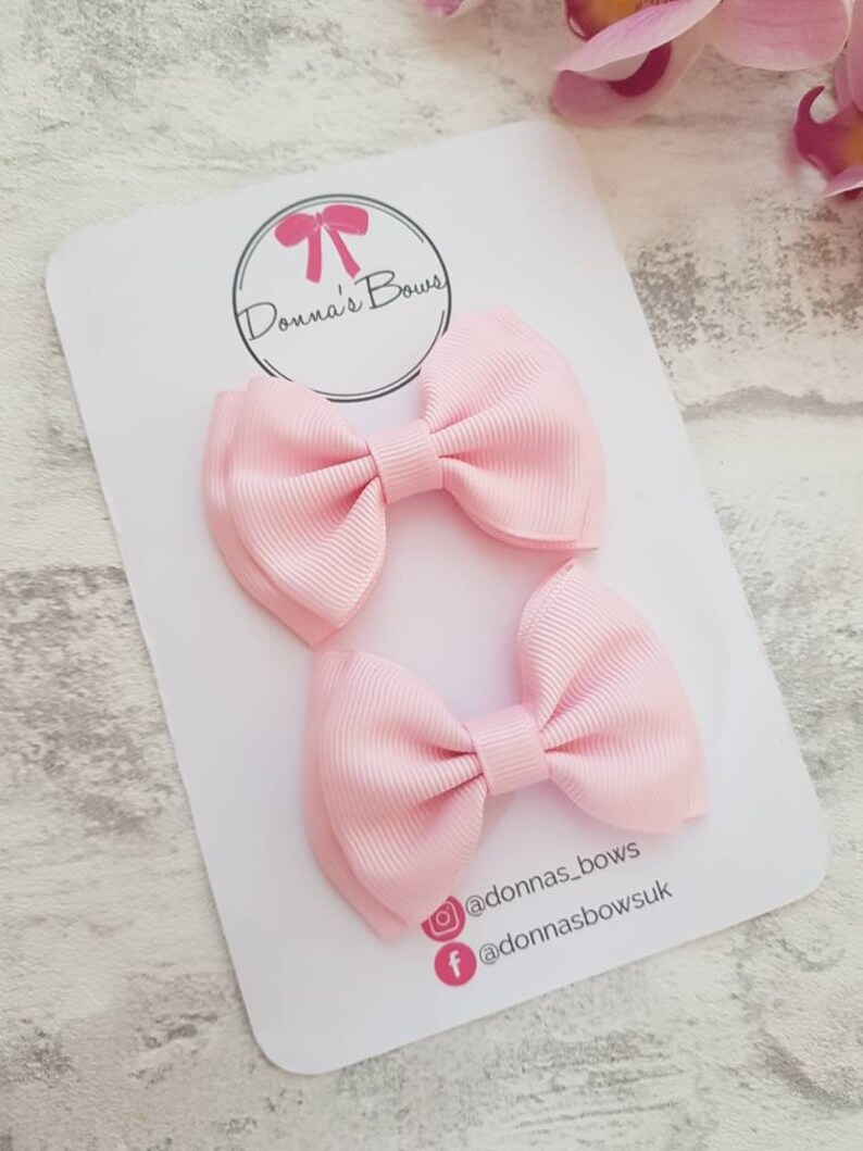 May include: Two light pink ribbon hair bows on a white card. The card has a circular logo with a pink bow and the text "Donna's Bows". Social media handles are also visible. The bows are made of a ribbed fabric.