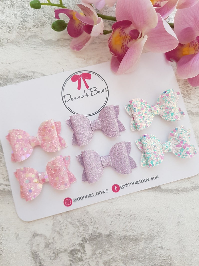 Peut inclure: Six n&oelig;uds en paillettes pour cheveux en rose, violet et blanc sur un fond blanc. Les n&oelig;uds sont dispos&eacute;s en trois rang&eacute;es de deux. Le texte "Donna's Bows" est dans un cercle en haut de la carte. Les poign&eacute;es de m&eacute;dias sociaux "@donnas_bows" et "@donnasbows.uk" sont en bas de la carte.