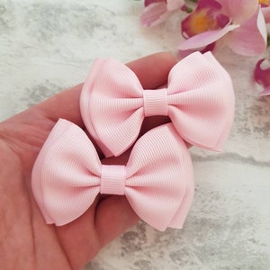 May include: Two light pink ribbon bows. The bows are made of a ribbed fabric and are tied in the center. The bows are held in a hand, with a blurred background of pink flowers and a gray and white surface.