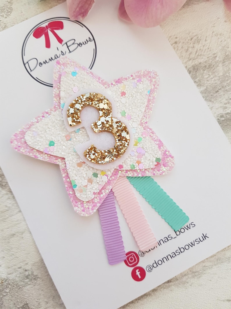 May include: A star-shaped hair clip with a glittery white and pink design, featuring a gold glitter number "3" in the center. Three pastel-colored ribbons in purple, pink, and teal are attached. The clip is on a white card with the text "Donna's Bows".
