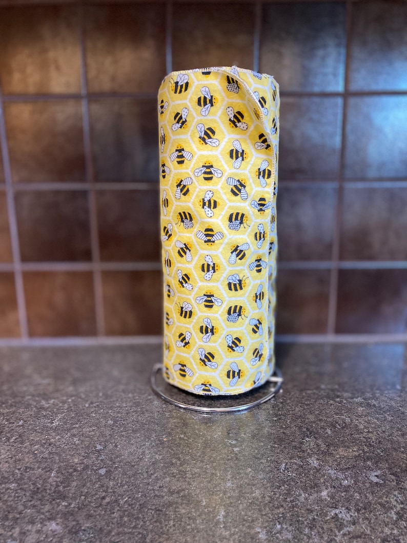 May include: A yellow and black reusable paper towel roll with a bee and honeycomb pattern. The roll is on a silver metal stand.