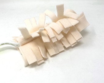 May include: A cream-colored duster head featuring multiple layers of thick, rectangular felt strips attached to a white plastic handle. The soft, fibrous material is arranged in a dense, layered pattern designed for cleaning surfaces.