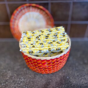 May include: A woven basket with a red and orange pattern holds a stack of yellow fabric squares with a bee pattern.