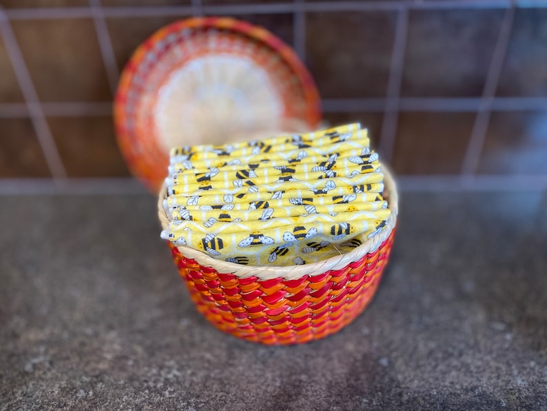 May include: A woven basket with a red and orange pattern holds a stack of yellow fabric squares with a bee pattern.