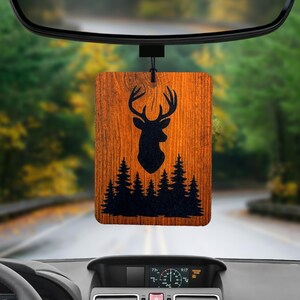 May include: A rectangular wooden car air freshener with a deer silhouette and forest design. The air freshener is brown with a black deer head and tree line. It hangs from a black string, visible through a car's rearview mirror.