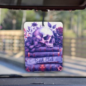 May include: A rectangular car air freshener featuring a purple skull atop a stack of books. The design includes purple flowers and the text "MORALLY GREY BOOK CLUB" in a decorative font. The air freshener hangs from a black string.