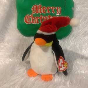Peut inclure: Un pingouin blanc et noir portant un bonnet de Père Noël rouge avec un pompon blanc. Le pingouin a un bec jaune et des yeux noirs. Le pingouin tient une étiquette Ty Beanie Baby rouge et blanche.
