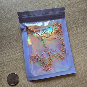 May include: A copper-colored wire headpiece with orange beads, resembling a branch, is displayed inside a lavender-edged iridescent bag. A U.S. quarter coin is next to the bag. The headpiece is likely for a special occasion.