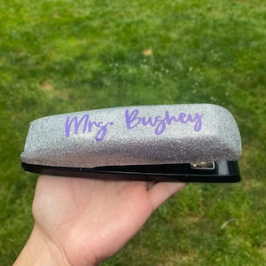 May include: A silver glitter stapler with the text "Mrs. Bughey" in purple script. The stapler is held in a hand against a green grass background. The stapler is black on the bottom.