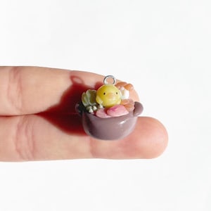 以下が含まれることがあります： 紫色の鍋に入った、小さな黄色のひよこの手作りのミニチュアチャーム。チャームは、ネックレスやブレスレットに取り付けるための銀色のループが付いています。チャームは人の指で持たれています。