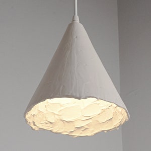 Handmade ceramic pendant lamp hanging above modern kitchen island with warm minimalist interior lighting. A white, cone-shaped pendant light fixture with a textured surface. The light is turned on and emits a warm glow.