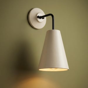 May include: A wall-mounted light fixture featuring a circular off-white base, a black metal L-shaped arm, and a conical off-white shade. The lamp emits a warm glow from the bottom opening, casting light downward against the wall surface.