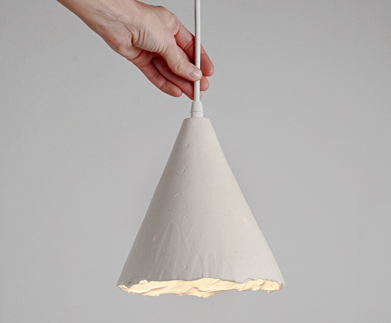 Rustic handmade ceramic hanging lamp designed for contemporary kitchen island and dining space. A white ceramic pendant light with a cone-shaped shade. The shade has a textured, wavy edge.