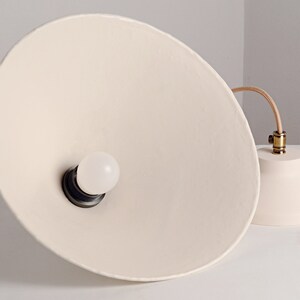 May include: A white ceramic pendant light with a round, white light bulb. The light fixture has a simple, modern design and is suspended from a brown cord.