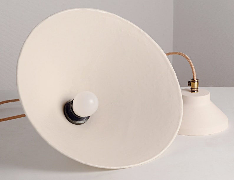 May include: A white ceramic pendant light with a round, white light bulb. The light fixture has a simple, modern design and is suspended from a brown cord.