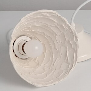 Close-up of artisan ceramic pendant light with natural texture and minimalist modern design. White ceramic pendant light fixture with a textured, wavy shade and a white bulb socket.