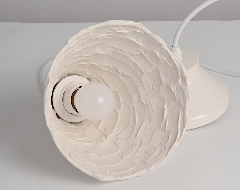 Close-up of artisan ceramic pendant light with natural texture and minimalist modern design. White ceramic pendant light fixture with a textured, wavy shade and a white bulb socket.