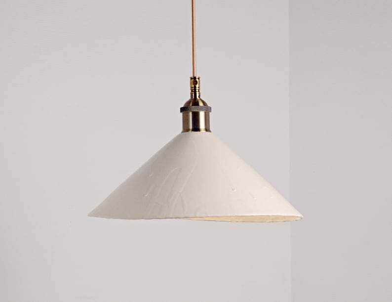 May include: A white ceramic pendant light with a brass fixture. The shade is a cone shape with a slightly uneven edge.