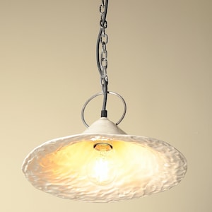 Scandinavian style ceramic pendant lamp with minimalist silhouette to hanfing above the kitchen island. White ceramic pendant light fixture with a textured, wavy surface. The light is suspended from a chain and has a single light bulb.