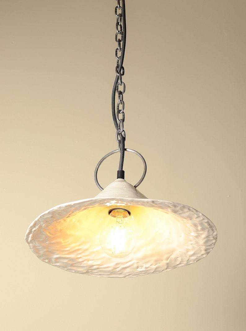 Scandinavian style ceramic pendant lamp with minimalist silhouette to hanfing above the kitchen island. White ceramic pendant light fixture with a textured, wavy surface. The light is suspended from a chain and has a single light bulb.