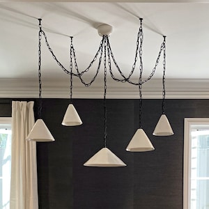 Customisation options. Contemporary handmade ceiling pendant light in ceramic material designed for kitchen island decor. A modern chandelier with six white cone-shaped shades hanging from black chains.