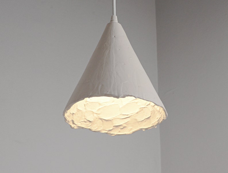 Handmade ceramic pendant lamp hanging above modern kitchen island with warm minimalist interior lighting. A white, cone-shaped pendant light fixture with a textured surface. The light is turned on and emits a warm glow.