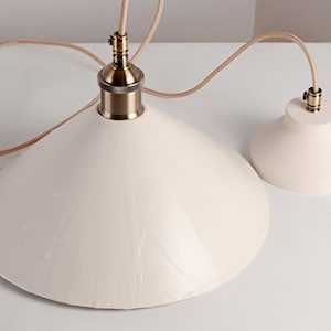 May include: Two white ceramic pendant light fixtures with brass accents and brown cords. The larger fixture has a wide, cone-shaped shade. The smaller fixture has a smaller, round shade.